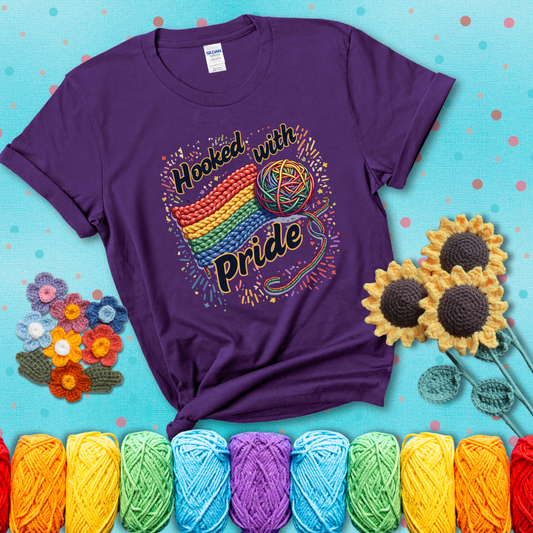 A rainbow-striped crochet swatch paired with a multicolor yarn ball and bold “Hooked With Pride” lettering. Confetti surrounds the vibrant scene showing a purple tee with a teal sunflower background