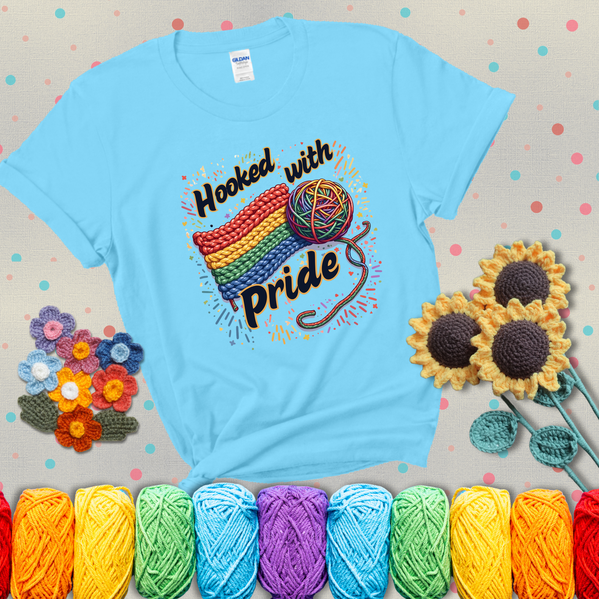 A rainbow-striped crochet swatch paired with a multicolor yarn ball and bold “Hooked With Pride” lettering. Confetti surrounds the vibrant scene showing a light blue tee with grey sunflower background