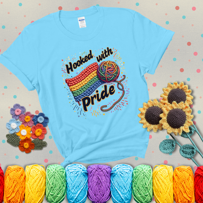 A rainbow-striped crochet swatch paired with a multicolor yarn ball and bold “Hooked With Pride” lettering. Confetti surrounds the vibrant scene showing a light blue tee with grey sunflower background