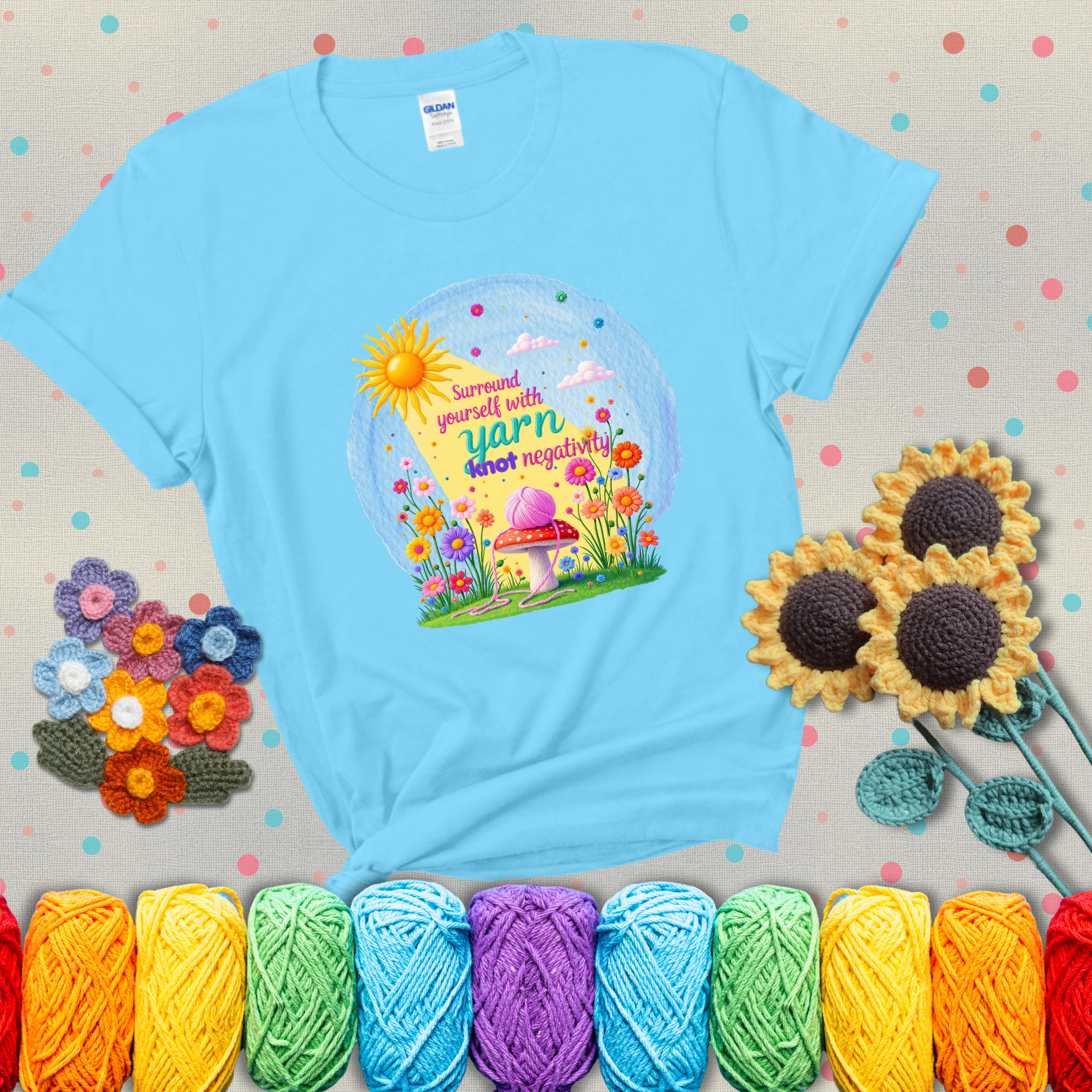 A happy spring garden scene with a sunbeam, flowers, and a pink yarn ball on a mushroom, with the quote "Surround yourself with yarn, knot negativity," showing a light blue tee with grey sunflower background