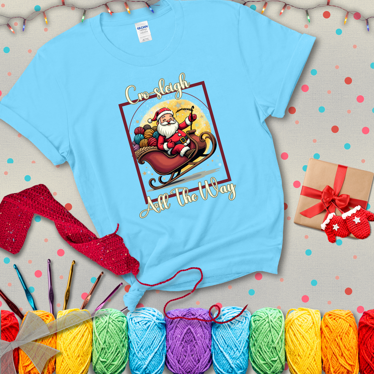 Jolly Santa rides a sleigh overflowing with colorful yarn balls, surrounded by sparkles and holiday cheer. Text reads “Cro-sleigh All the Way" showing a light blue tee with a festive Christmas background