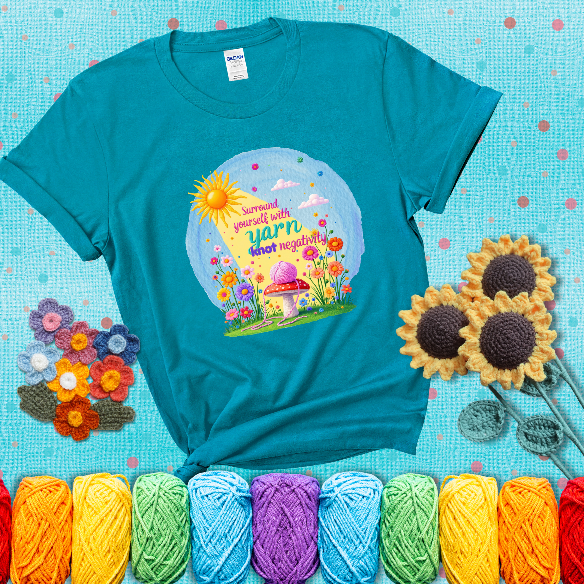 A happy spring garden scene with a sunbeam, flowers, and a pink yarn ball on a mushroom, with the quote "Surround yourself with yarn, knot negativity," showing a blue-green tee with a teal sunflower background