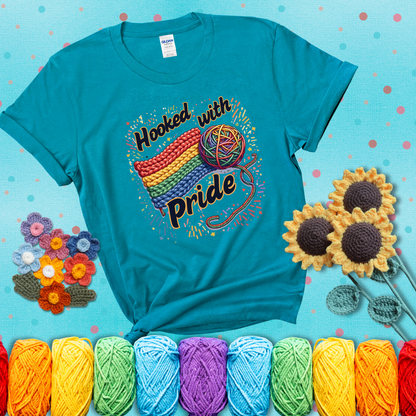 A rainbow-striped crochet swatch paired with a multicolor yarn ball and bold “Hooked With Pride” lettering. Confetti surrounds the vibrant scene showing a blue-green tee with a teal sunflower background