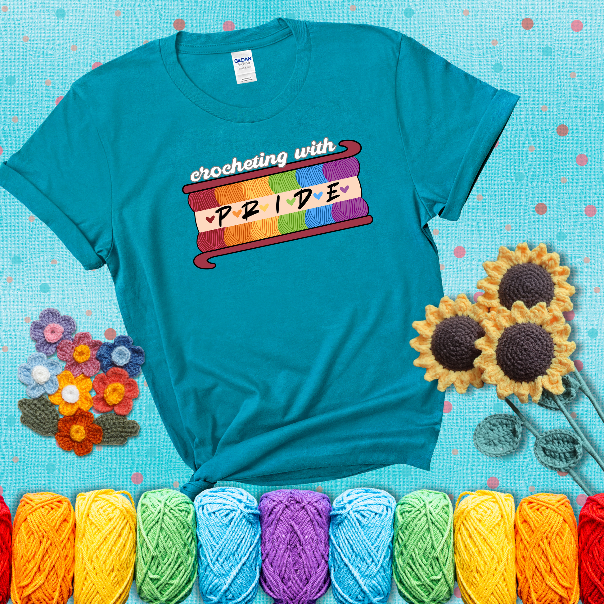 Yarn skeins in rainbow colors stacked in a hook shape. Text reads “Crocheting With Pride” showing a blue-green tee with a teal sunflower background