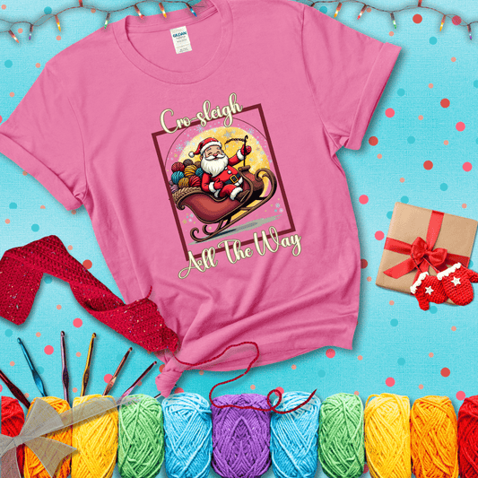 Jolly Santa rides a sleigh overflowing with colorful yarn balls, surrounded by sparkles and holiday cheer. Text reads “Cro-sleigh All the Way" showing a pink tee with a festive Christmas background