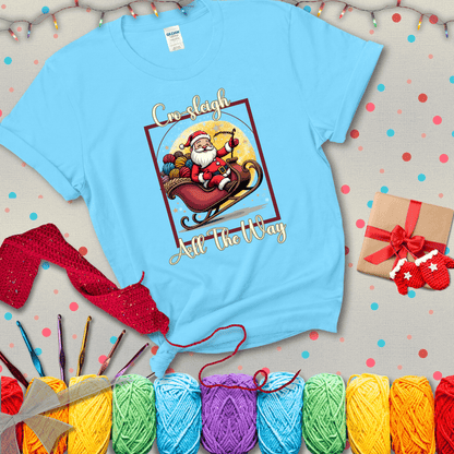 Jolly Santa rides a sleigh overflowing with colorful yarn balls, surrounded by sparkles and holiday cheer. Text reads “Cro-sleigh All the Way" showing a light blue tee with a festive Christmas background