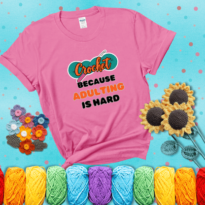 A bold design with the phrase “Crochet Because Adulting Is Hard” featuring a green skein of yarn with a pink crochet hook. The word “Crochet” is in a script font, while “Adulting is hard” is in chunky textured letters showing a pink tee with a teal sunflower background