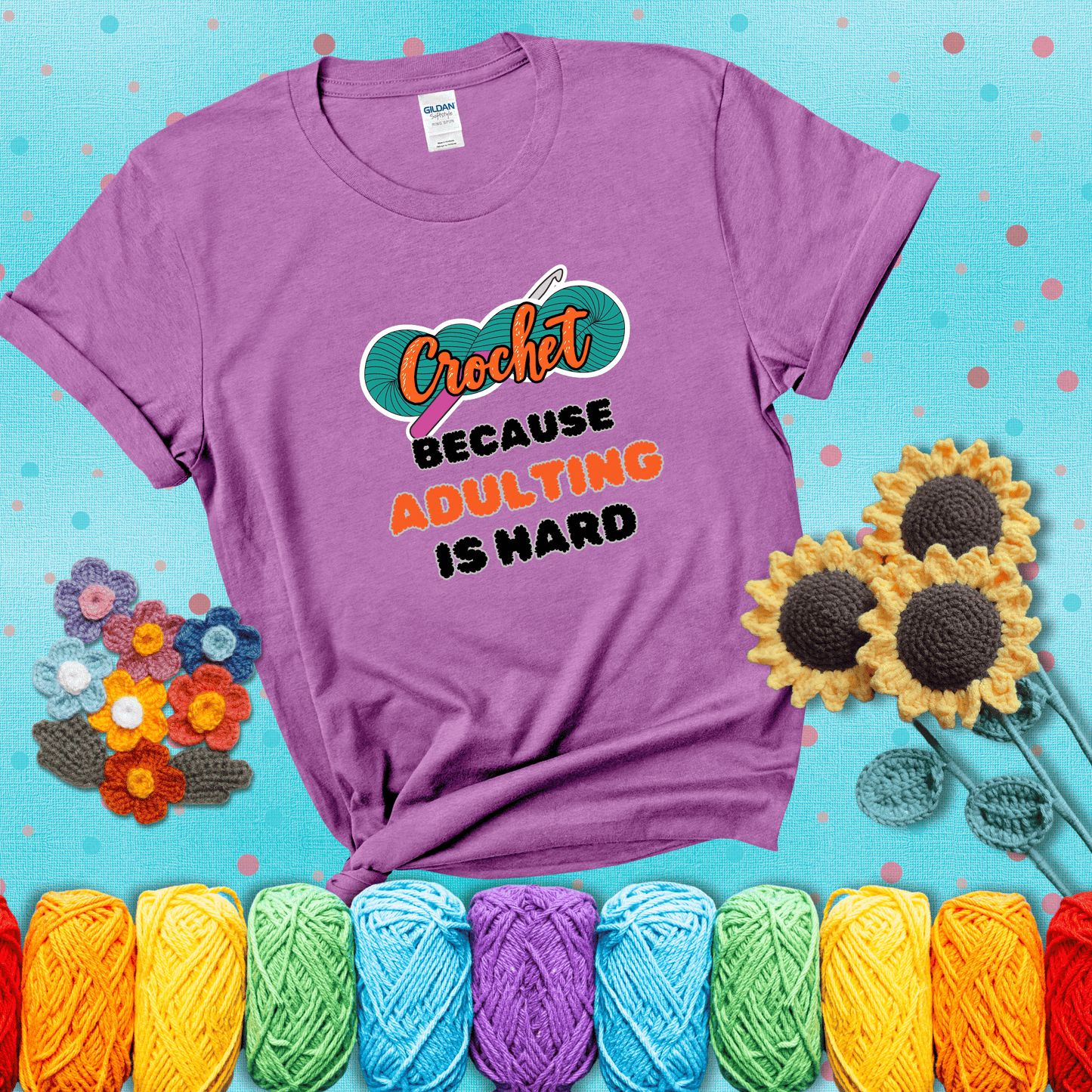 A bold design with the phrase “Crochet Because Adulting Is Hard” featuring a green skein of yarn with a pink crochet hook. The word “Crochet” is in a script font, while “Adulting is hard” is in chunky textured letters showing a light purple tee with a teal sunflower background