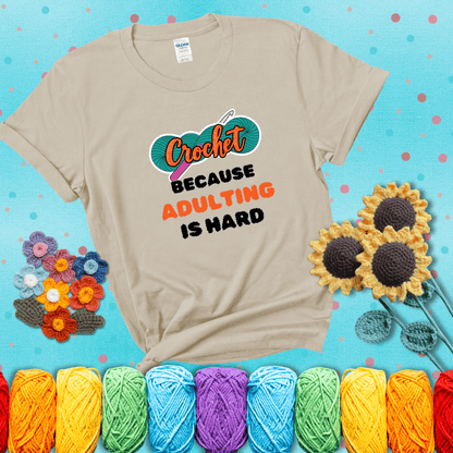 A bold design with the phrase “Crochet Because Adulting Is Hard” featuring a green skein of yarn with a pink crochet hook. The word “Crochet” is in a script font, while “Adulting is hard” is in chunky textured letters showing a beige tee with a teal sunflower background