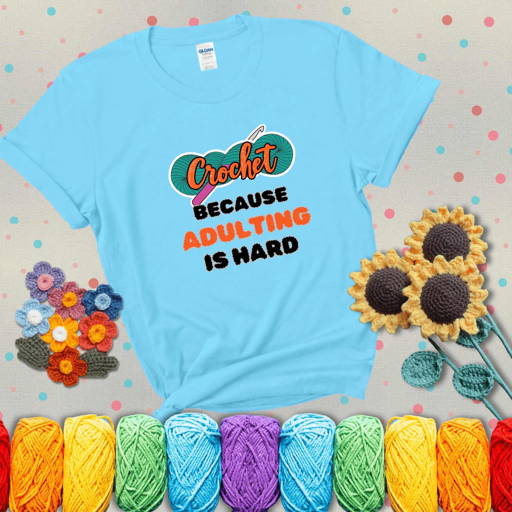 A bold design with the phrase “Crochet Because Adulting Is Hard” featuring a green skein of yarn with a pink crochet hook. The word “Crochet” is in a script font, while “Adulting is hard” is in chunky textured letters showing a light blue tee with grey sunflower background