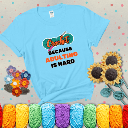 A bold design with the phrase “Crochet Because Adulting Is Hard” featuring a green skein of yarn with a pink crochet hook. The word “Crochet” is in a script font, while “Adulting is hard” is in chunky textured letters showing a light blue tee with grey sunflower background