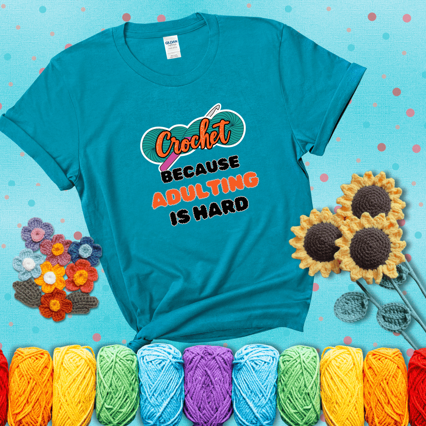 A bold design with the phrase “Crochet Because Adulting Is Hard” featuring a green skein of yarn with a pink crochet hook. The word “Crochet” is in a script font, while “Adulting is hard” is in chunky textured letters showing a blue-green tee with a teal sunflower background