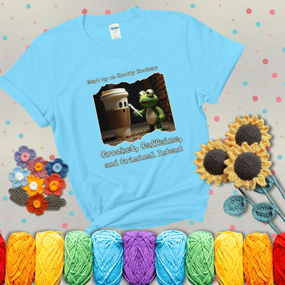 A crocheted frog holding a crochet hook confronts a surprised coffee cup in a dim alleyway with text: "Next up on Knotty Hooker: Crochet, Caffeine, and Criminal Intent," showing a light blue tee with grey sunflower background