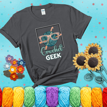 A teal yarn ball with crisscrossed hooks wears oversized nerdy glasses with the phrase “Crochet Geek” in bold, fun text below, showing a dark grey tee with a teal sunflower background