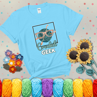 A teal yarn ball with crisscrossed hooks wears oversized nerdy glasses with the phrase “Crochet Geek” in bold, fun text below, showing a light blue tee with grey sunflower background
