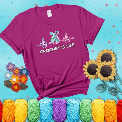 A heartbeat line with a colorful yarn ball and crochet hook in the center, with the words “Crochet is Life”, showing a magenta tee with a teal sunflower background
