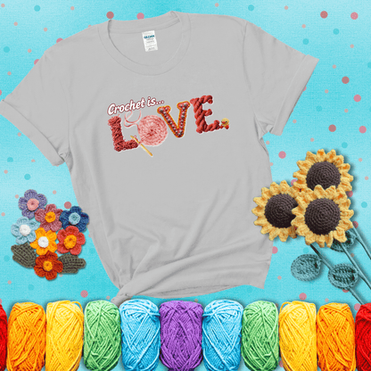 The word "LOVE" spelled with different crochet textures and yarn in red tones, with the phrase “Crochet is…” above in script. showing a light blue tee with grey sunflower background