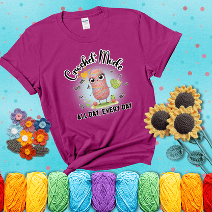A kawaii-style rainbow yarn character surrounded by crochet stitch abbreviations and holding a yarn heart, with text “Crochet Mode All Day, Every Day"; showing a magenta tee with a teal sunflower background