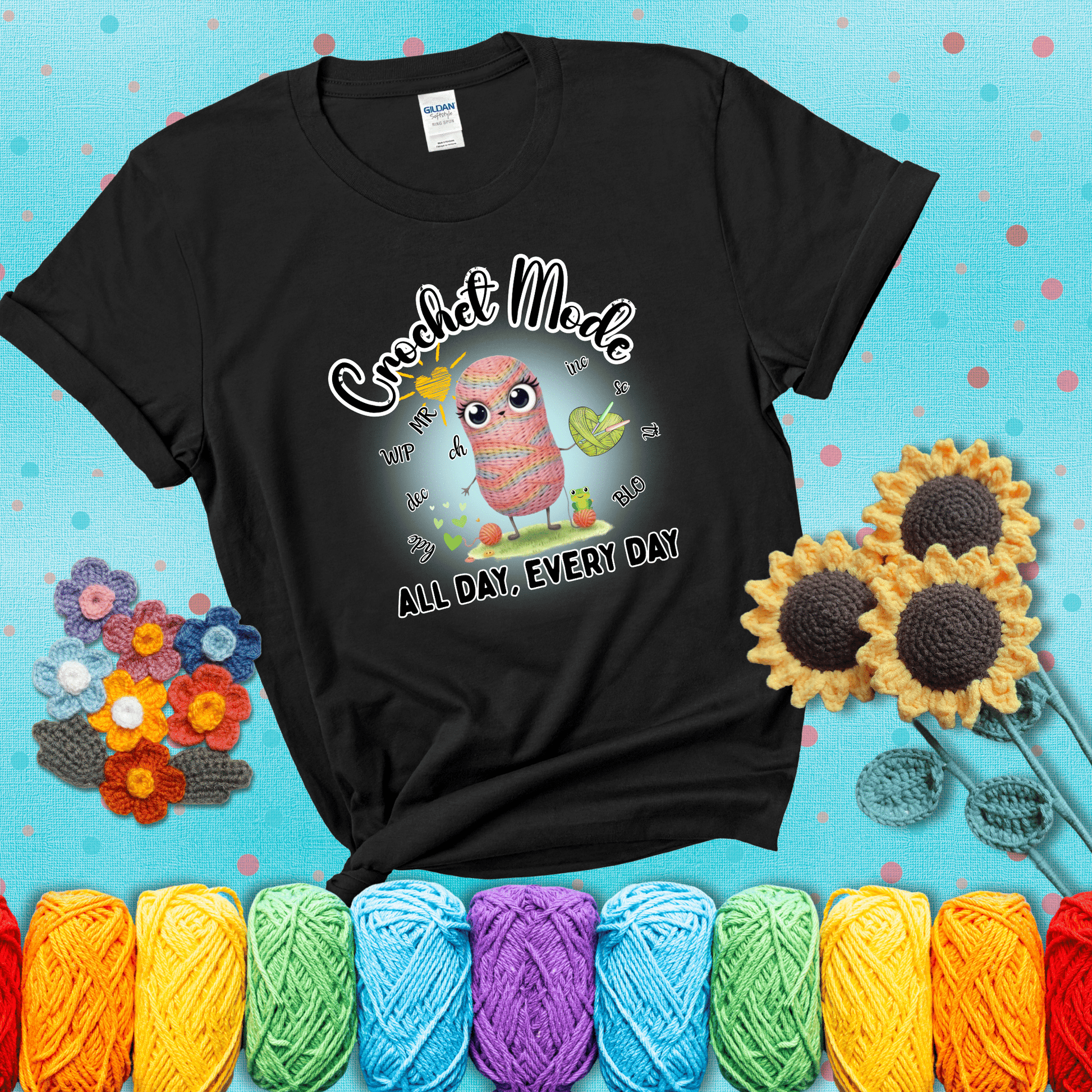 A kawaii-style rainbow yarn character surrounded by crochet stitch abbreviations and holding a yarn heart, with text “Crochet Mode All Day, Every Day"; showing a black tee with a teal sunflower background