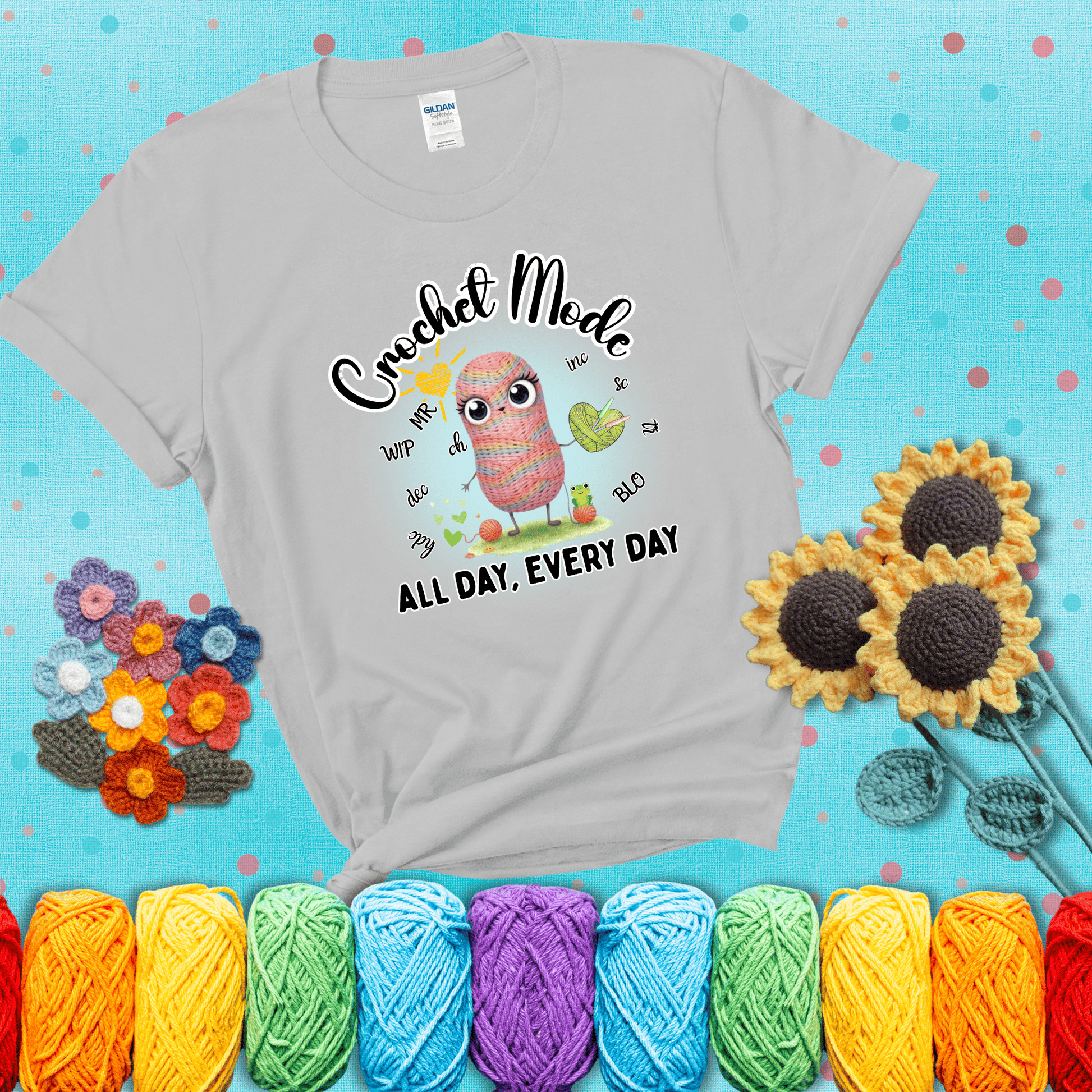 A kawaii-style rainbow yarn character surrounded by crochet stitch abbreviations and holding a yarn heart, with text “Crochet Mode All Day, Every Day"; showing a light grey tee with a teal sunflower background