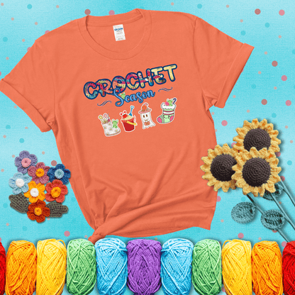 The words “Crochet Season” with cute seasonal icons below: a spring boot with hooks, summer bucket with crab, fall ghost with pumpkin hat, and winter stocking - all with tiny frogs; showing a light orange tee with a teal sunflower background