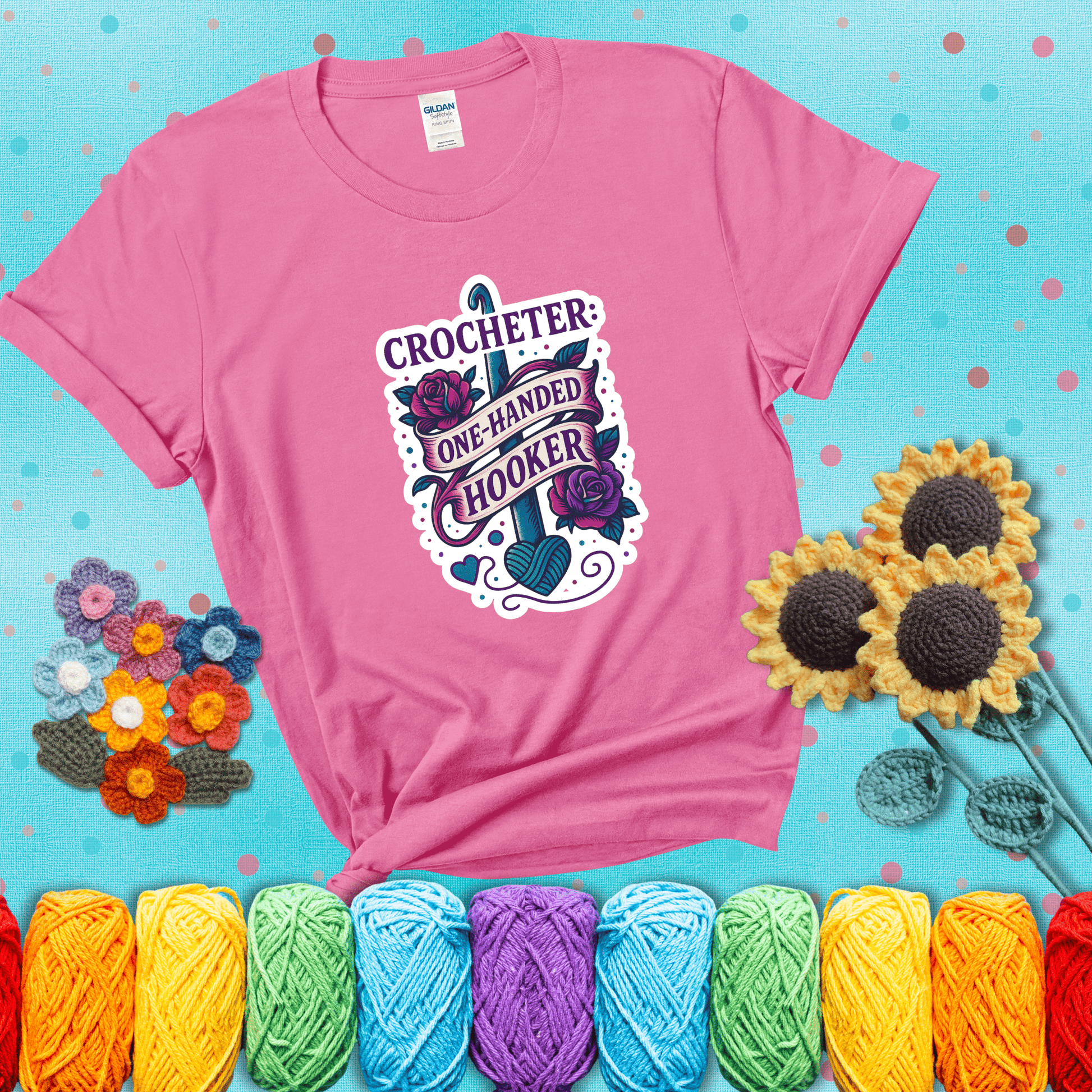 Tattoo-style design with a crochet hook, roses, and banner that says "Crocheter: One-Handed Hooker" showing a pink tee with a teal sunflower background