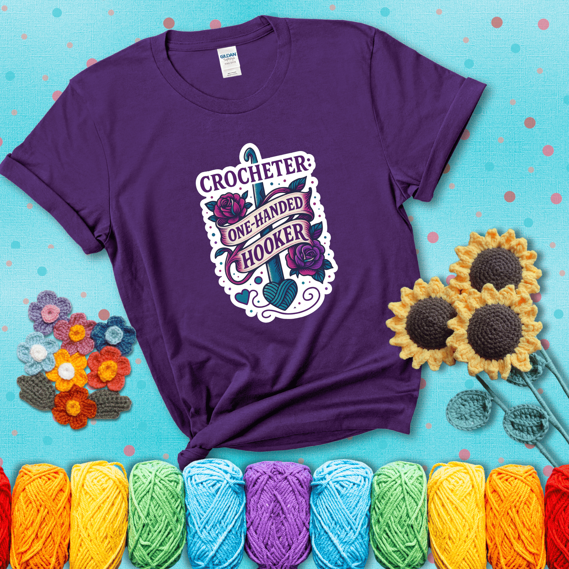 Tattoo-style design with a crochet hook, roses, and banner that says "Crocheter: One-Handed Hooker" showing a purple tee with a teal sunflower background