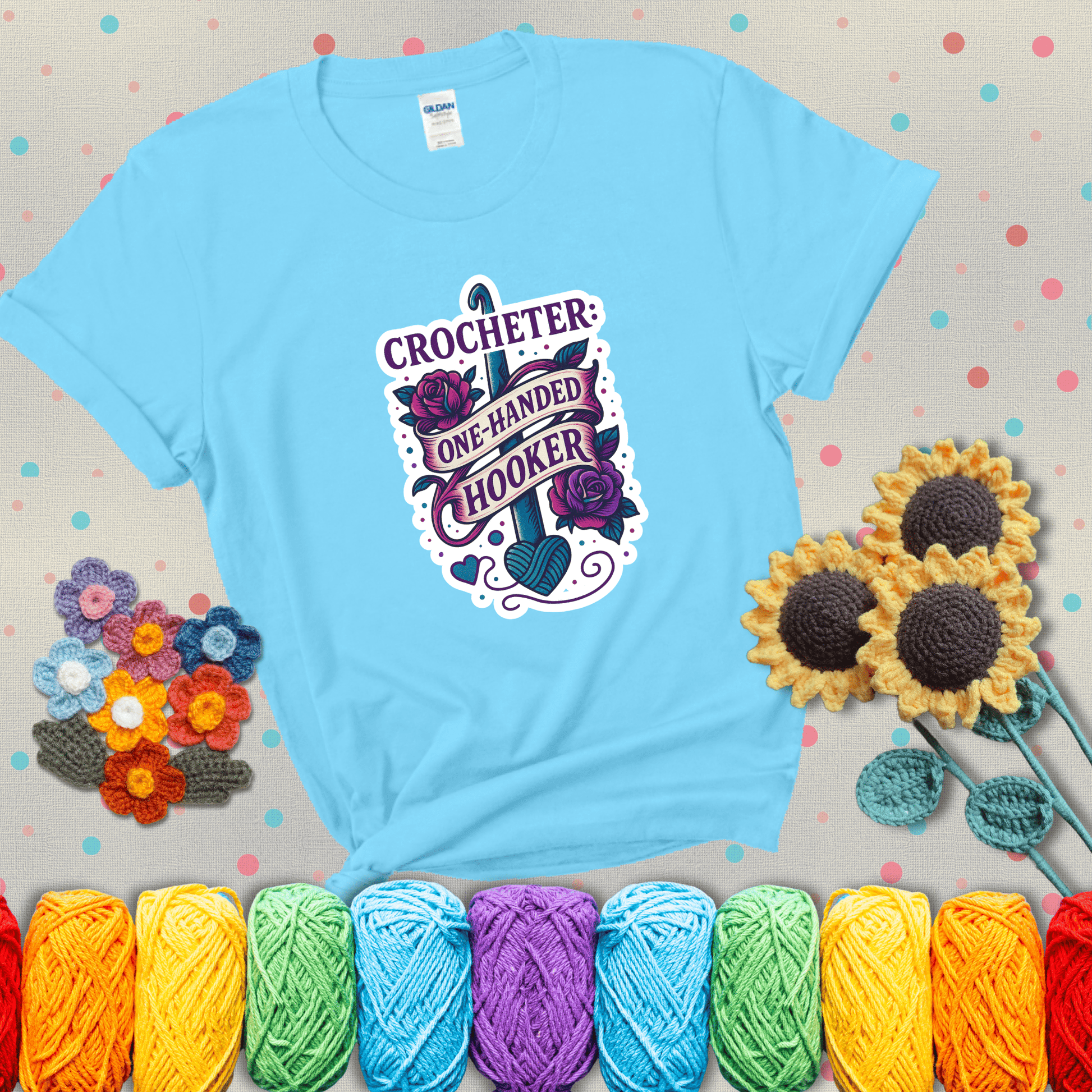Tattoo-style design with a crochet hook, roses, and banner that says "Crocheter: One-Handed Hooker" showing a light blue tee with grey sunflower background