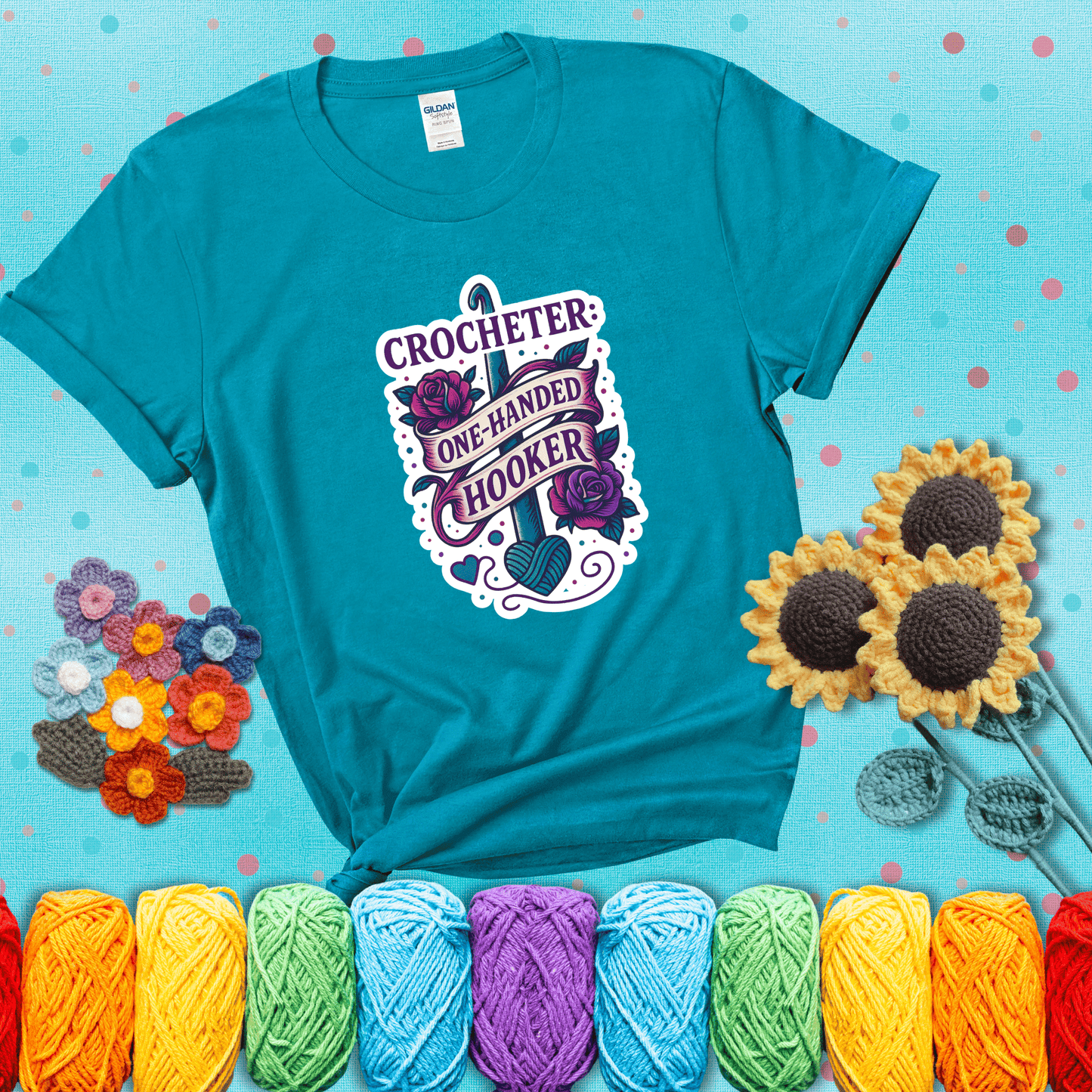 Tattoo-style design with a crochet hook, roses, and banner that says "Crocheter: One-Handed Hooker" showing a blue-green tee with a teal sunflower background