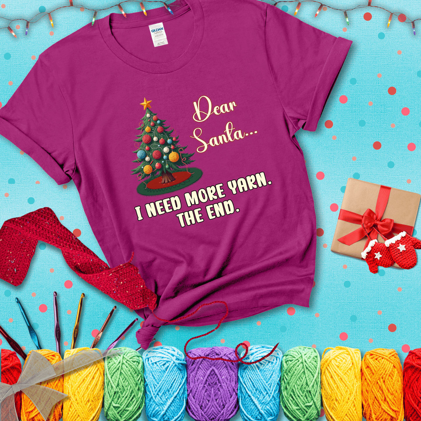 Christmas tree decorated with yarn ball ornaments and a stitched message that reads “Dear Santa… I Need More Yarn. The End.” Whimsical holiday yarn-lover shirt, showing a magenta tee with a festive Christmas background