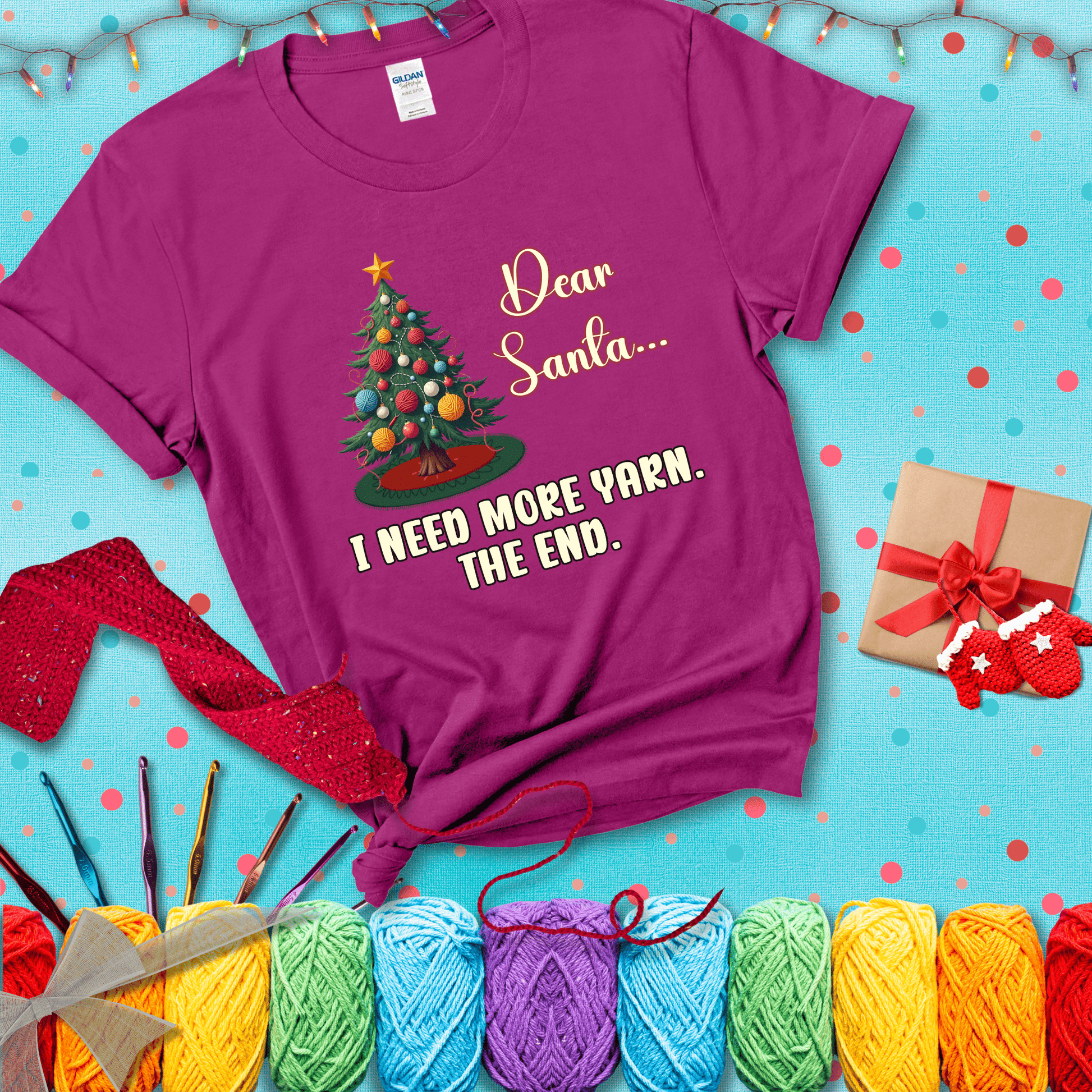 Christmas tree decorated with yarn ball ornaments and a stitched message that reads “Dear Santa… I Need More Yarn. The End.” Whimsical holiday yarn-lover shirt, showing a magenta tee with a festive Christmas background