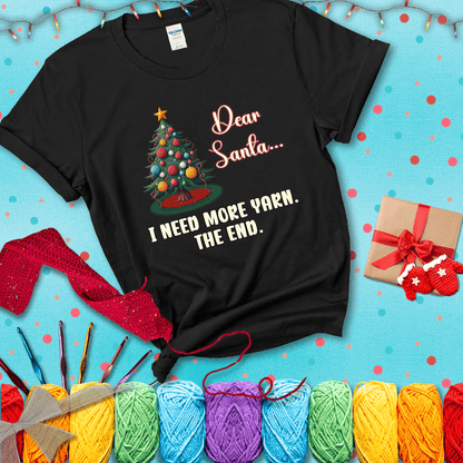 Christmas tree decorated with yarn ball ornaments and a stitched message that reads “Dear Santa… I Need More Yarn. The End.” Whimsical holiday yarn-lover shirt, showing a black tee with a festive Christmas background