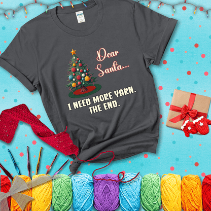 Christmas tree decorated with yarn ball ornaments and a stitched message that reads “Dear Santa… I Need More Yarn. The End.” Whimsical holiday yarn-lover shirt, showing a dark grey tee with a festive Christmas background