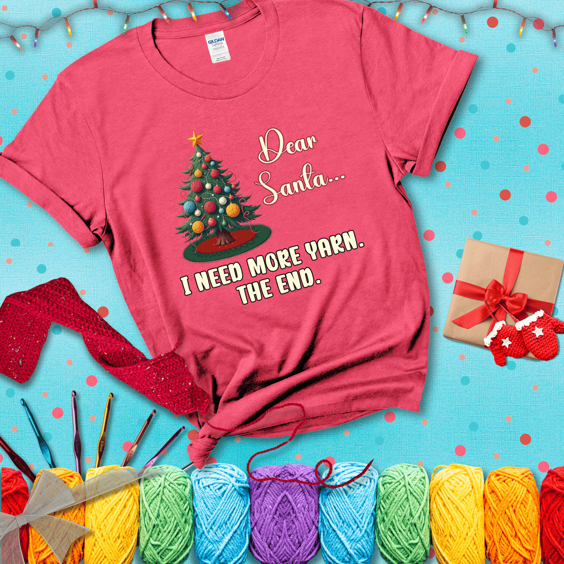 Christmas tree decorated with yarn ball ornaments and a stitched message that reads “Dear Santa… I Need More Yarn. The End.” Whimsical holiday yarn-lover shirt, showing a light red tee with a festive Christmas background