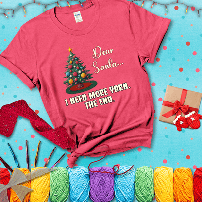 Christmas tree decorated with yarn ball ornaments and a stitched message that reads “Dear Santa… I Need More Yarn. The End.” Whimsical holiday yarn-lover shirt, showing a light red tee with a festive Christmas background