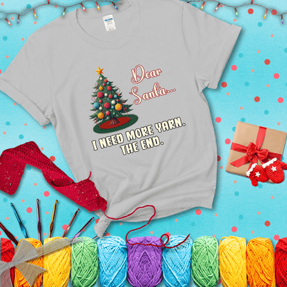 Christmas tree decorated with yarn ball ornaments and a stitched message that reads “Dear Santa… I Need More Yarn. The End.” Whimsical holiday yarn-lover shirt, showing a light grey tee with a festive Christmas background