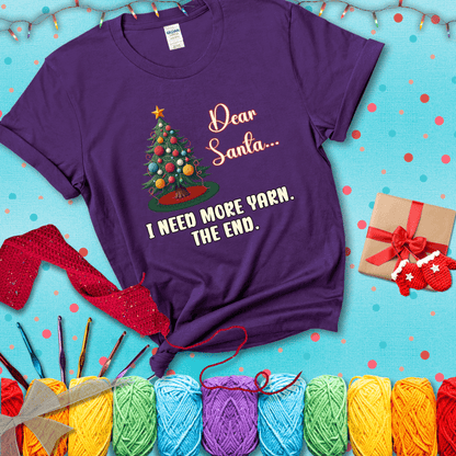 Christmas tree decorated with yarn ball ornaments and a stitched message that reads “Dear Santa… I Need More Yarn. The End.” Whimsical holiday yarn-lover shirt, showing a purple tee with a festive Christmas background