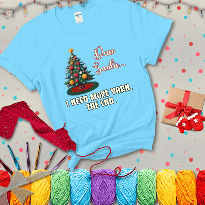 Christmas tree decorated with yarn ball ornaments and a stitched message that reads “Dear Santa… I Need More Yarn. The End.” Whimsical holiday yarn-lover shirt, showing a light blue tee with a festive Christmas background