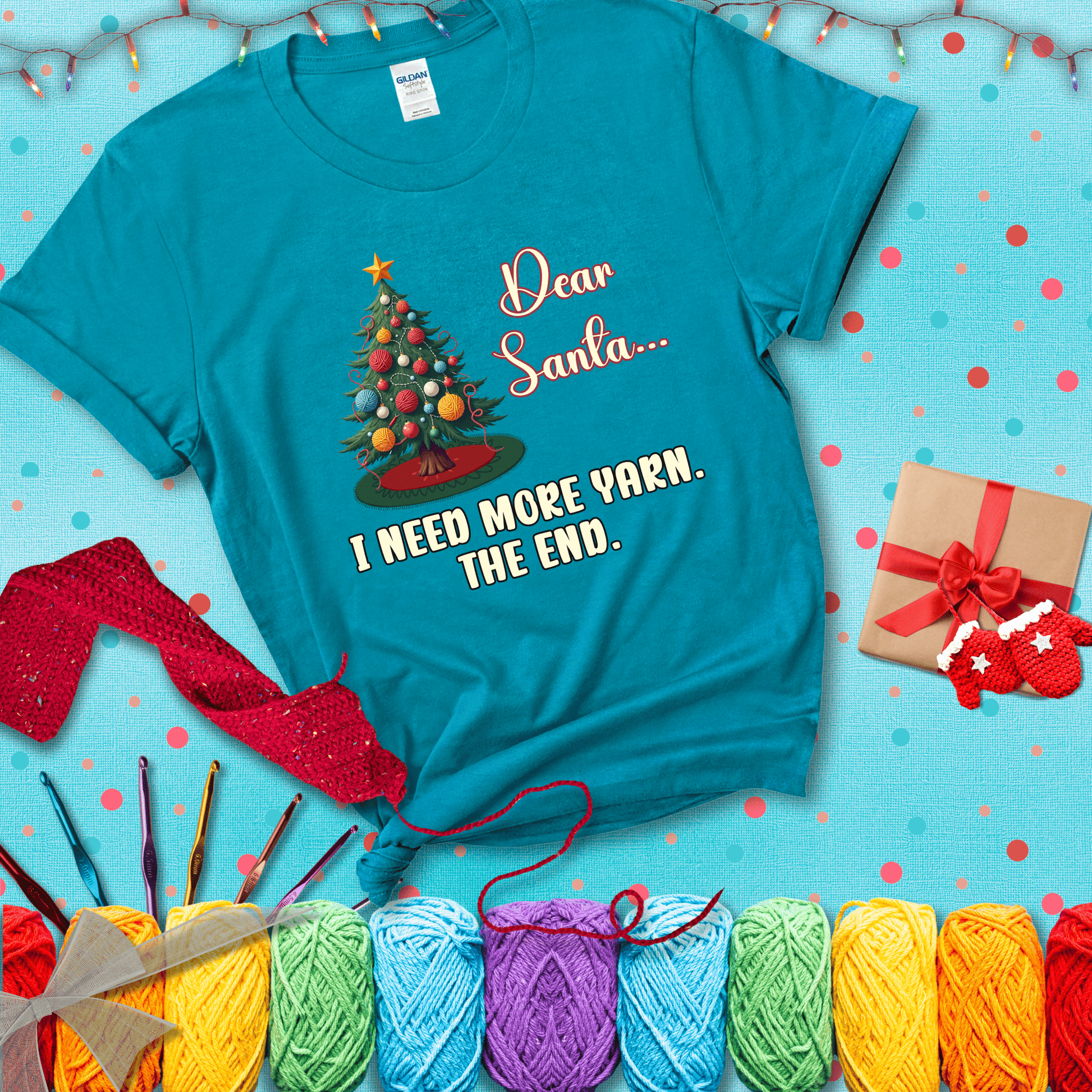 Christmas tree decorated with yarn ball ornaments and a stitched message that reads “Dear Santa… I Need More Yarn. The End.” Whimsical holiday yarn-lover shirt, showing a blue-green tee with a festive Christmas background