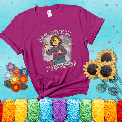 Annoyed expression with yarn and a hand held up to the viewer. Text reads: “Don’t Talk to Me, I’m Counting.” A relatable tee for focused crocheters and knitters alike, showing a magenta tee with a teal sunflower background