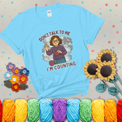 Annoyed expression with yarn and a hand held up to the viewer. Text reads: “Don’t Talk to Me, I’m Counting.” A relatable tee for focused crocheters and knitters alike, showing a light blue tee with grey sunflower background