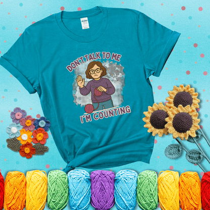 Annoyed expression with yarn and a hand held up to the viewer. Text reads: “Don’t Talk to Me, I’m Counting.” A relatable tee for focused crocheters and knitters alike, showing a blue-green tee with a teal sunflower background