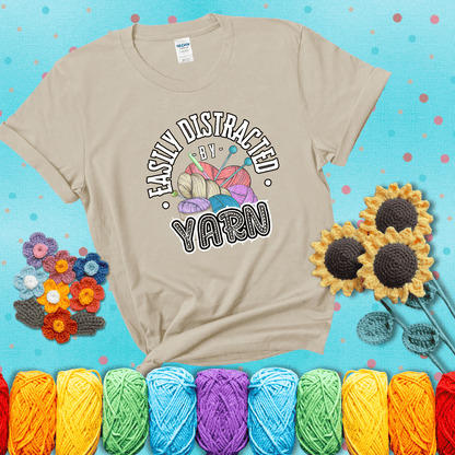 If yarn steals your focus every Design reads 'Easily Distracted By Yarn', perfect crochet gift for yarn lovers; showing a beige tee with a teal sunflower background