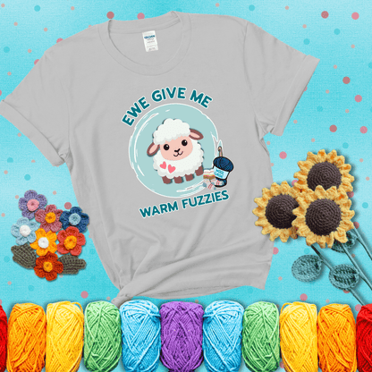 Cartoon sheep with pink hearts, wool socks, and yarn labeled “Wool Classic.” Text reads “Ewe Give Me Warm Fuzzies” showing a light grey tee with a teal sunflower background