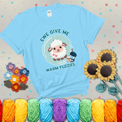 Cartoon sheep with pink hearts, wool socks, and yarn labeled “Wool Classic.” Text reads “Ewe Give Me Warm Fuzzies” showing a light blue tee with grey sunflower background