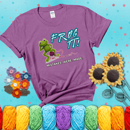 Green frog with yarn and heart-shaped loop. Text reads: “Frog It! Mistakes Were Made.” A playful crochet tee for anyone who's ever had to rip it back, showing a light purple tee with a teal sunflower background