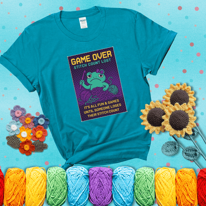 Pixel-style design of a grumpy yarn frog holding a hook, surrounded by yarn chaos, with the text “Game Over. Stitch Count Lost” showing a blue-green tee with a teal sunflower background