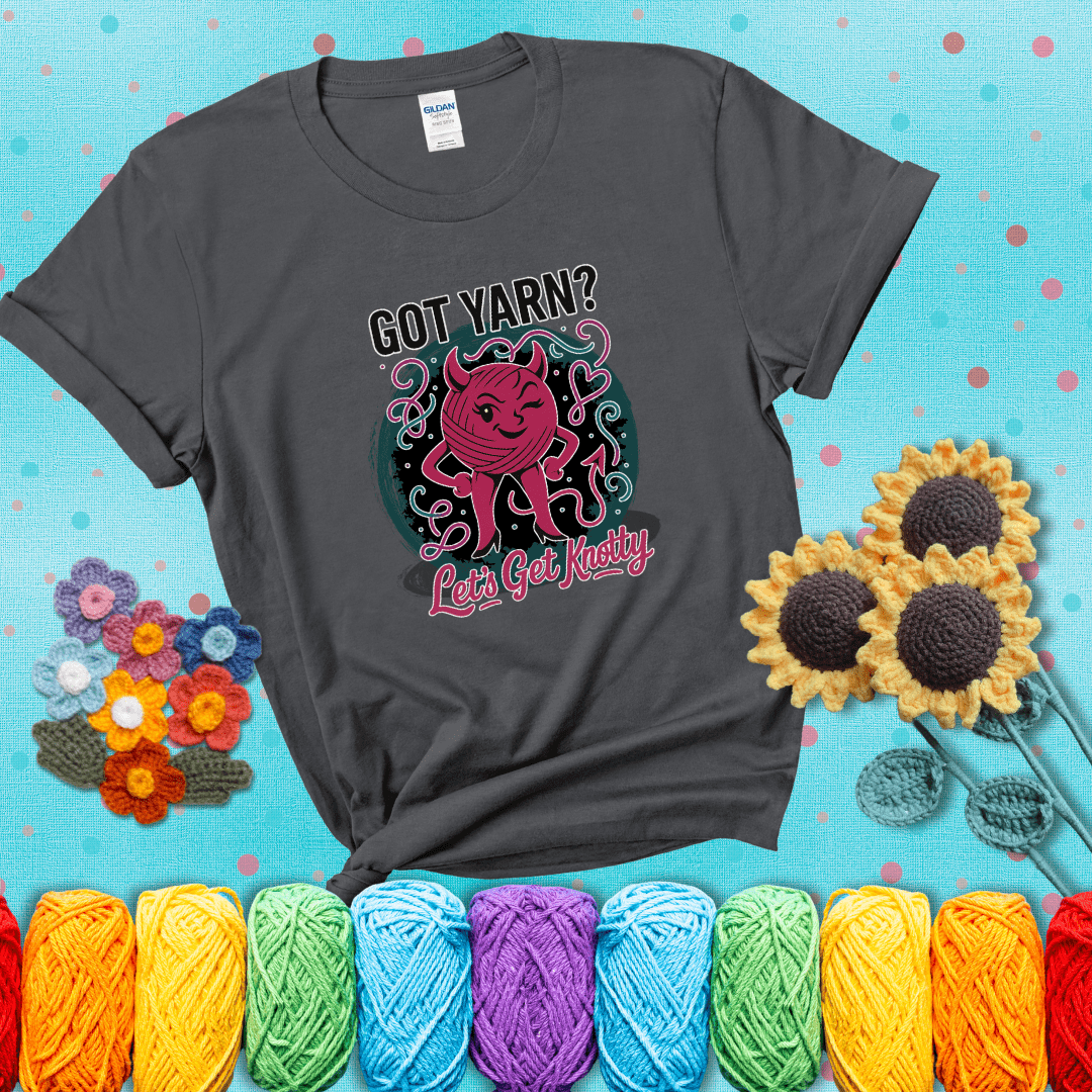 A cheeky pink yarn ball with devil horns, arms, and legs winks under the text “Got Yarn? Let’s Get Knotty,” showing a dark grey tee with a teal sunflower background
