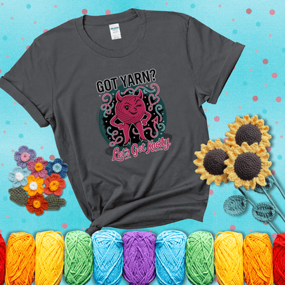 A cheeky pink yarn ball with devil horns, arms, and legs winks under the text “Got Yarn? Let’s Get Knotty,” showing a dark grey tee with a teal sunflower background
