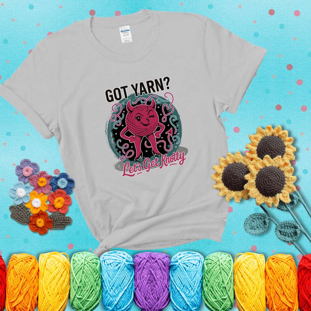 A cheeky pink yarn ball with devil horns, arms, and legs winks under the text “Got Yarn? Let’s Get Knotty,” showing a light grey tee with a teal sunflower background